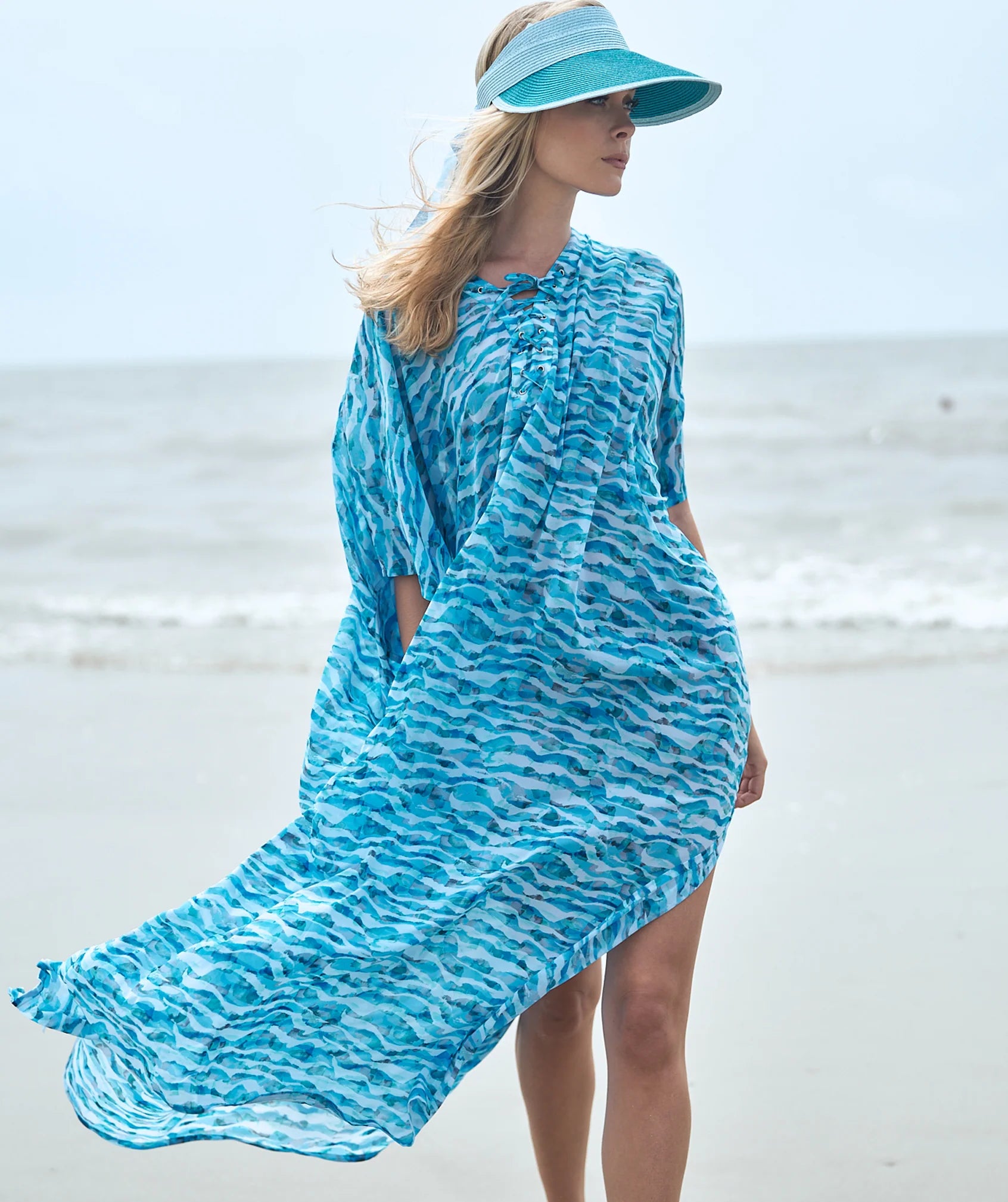 Woman in a blue patterned maxi length kaftan beach cover-up and visor standing on a beach.